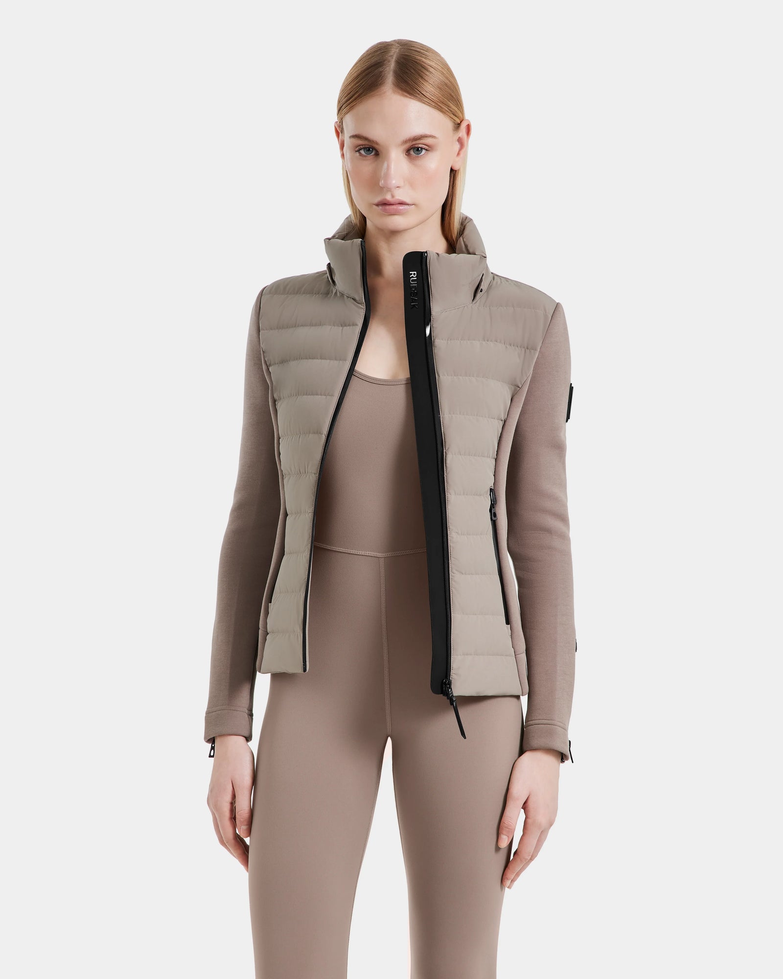 A woman with straight, light brown hair wears the Rudsak MABS - 8124825 puffer vest in Cement over a taupe, long-sleeve fitted jumpsuit, standing against a plain white background.