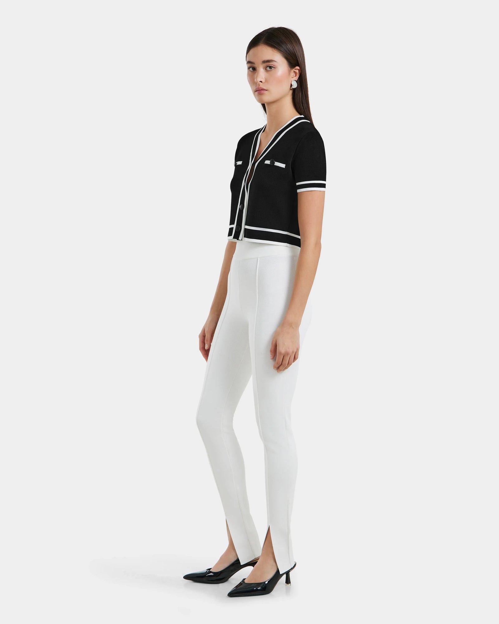 A woman stands against a white background wearing a short-sleeve black top with white trim, MATHILDA - 8524515 Iceland high-waisted front ankle slit leggings by Rudsak, and black low-heeled shoes.