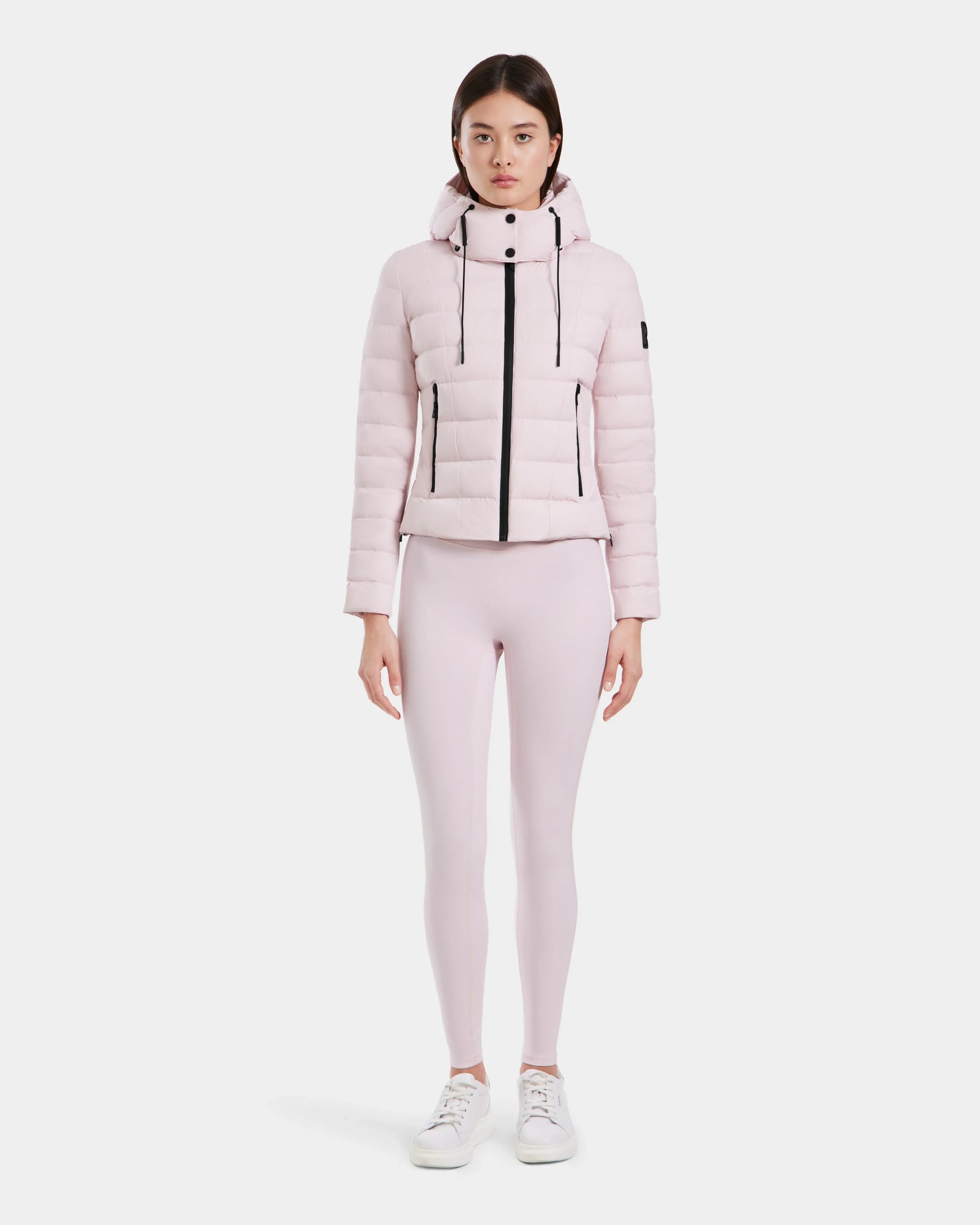 A woman faces forward, wearing the Rudsak MELISSA - 8125054 light pink puffer jacket with a hood, matching light pink leggings, and white sneakers against a plain white background.