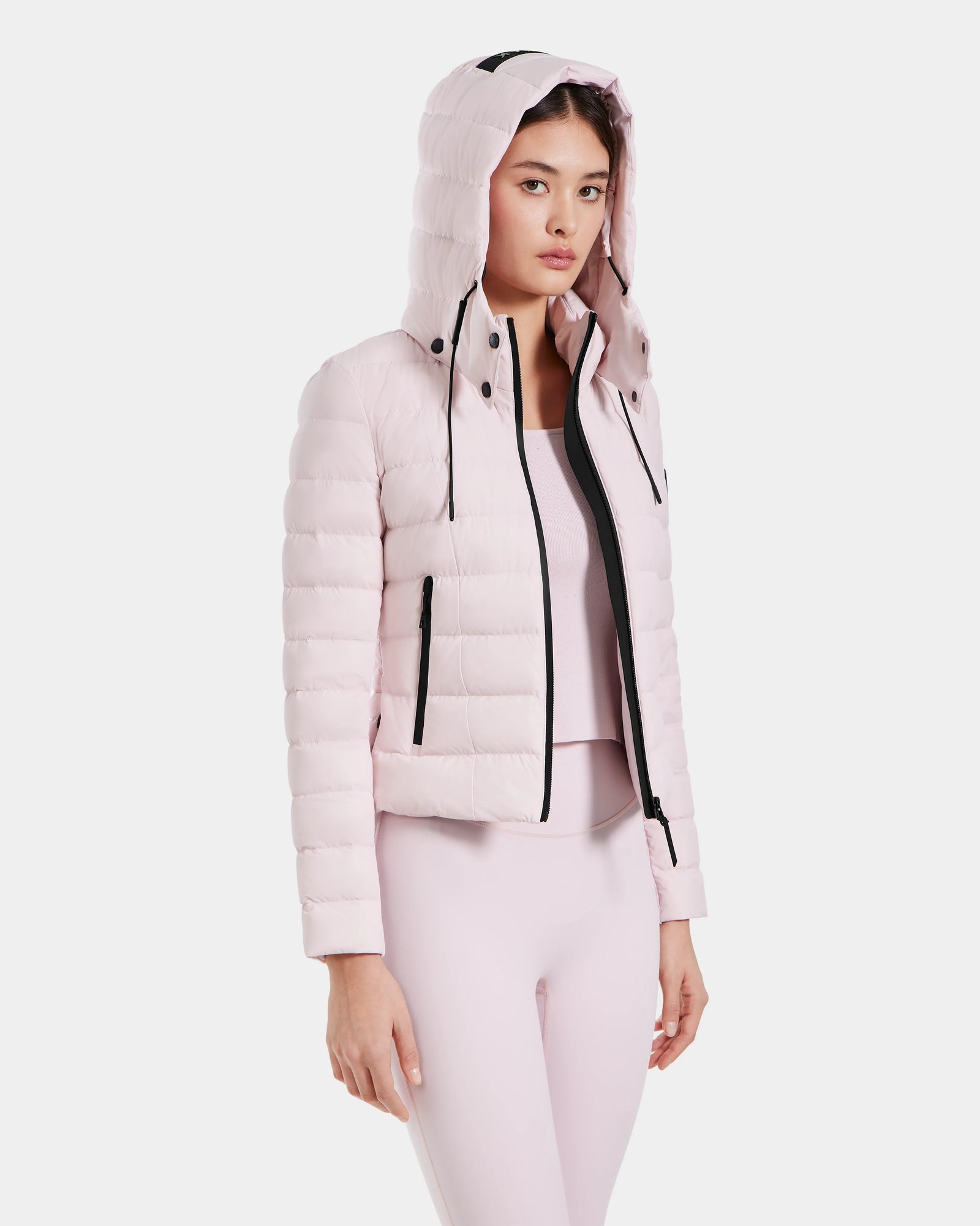 A woman in the Rudsak MELISSA - 8125054 Light Pink water-resistant puffer jacket with a hood, paired with matching light pink leggings and top, stands turned slightly to the side against a white background.