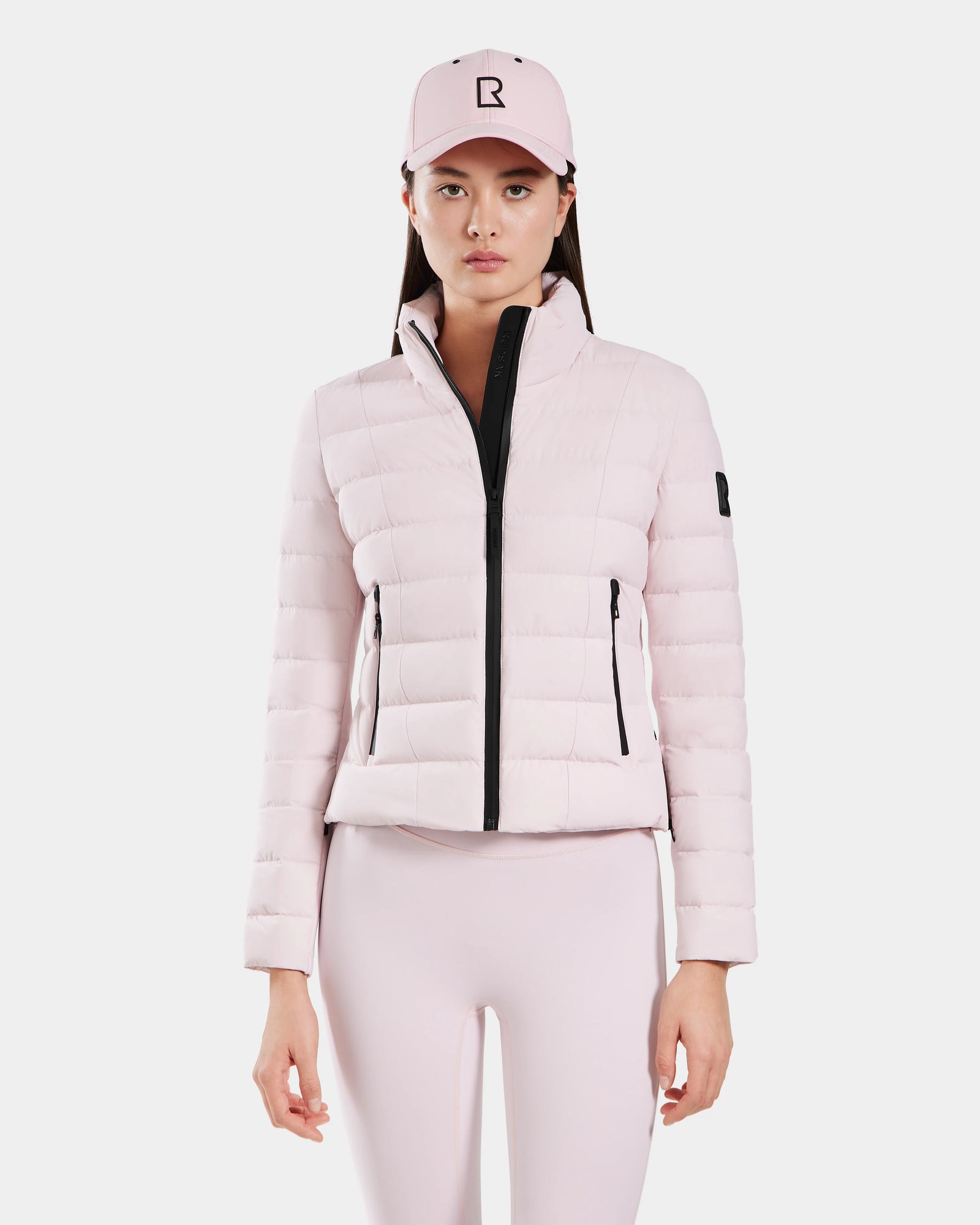 A woman models the Rudsak MELISSA - 8125054 Light Pink puffer jacket with a black front zipper and zippered pockets, paired with matching pink leggings and a baseball cap, against a plain white background.