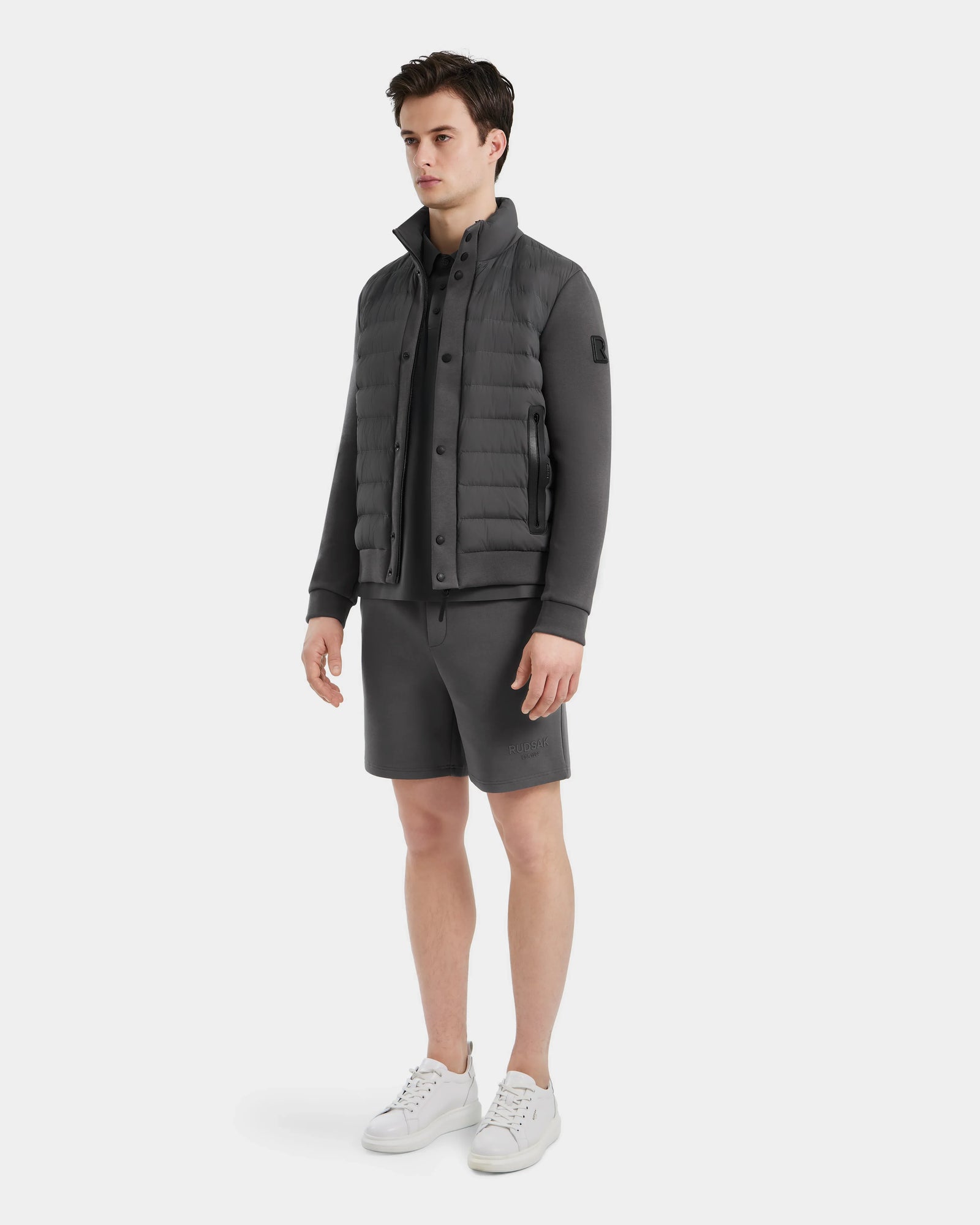 A man stands against a white background wearing the Rudsak RAGAB - 6124577 Grey hybrid jacket with lightweight down insulation, matching zip-up, grey shorts, and white sneakers—perfect for transitional weather. He looks slightly to the side, hands relaxed.