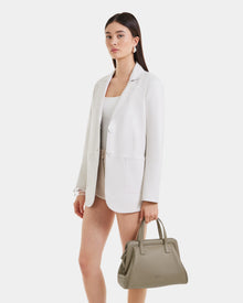 A woman with straight dark hair and earrings stands against a white background, wearing a white blazer and shorts. She holds the Rudsak TAULLA - 8325002 Almond everyday essential pebble leather satchel with a removable shoulder strap.
