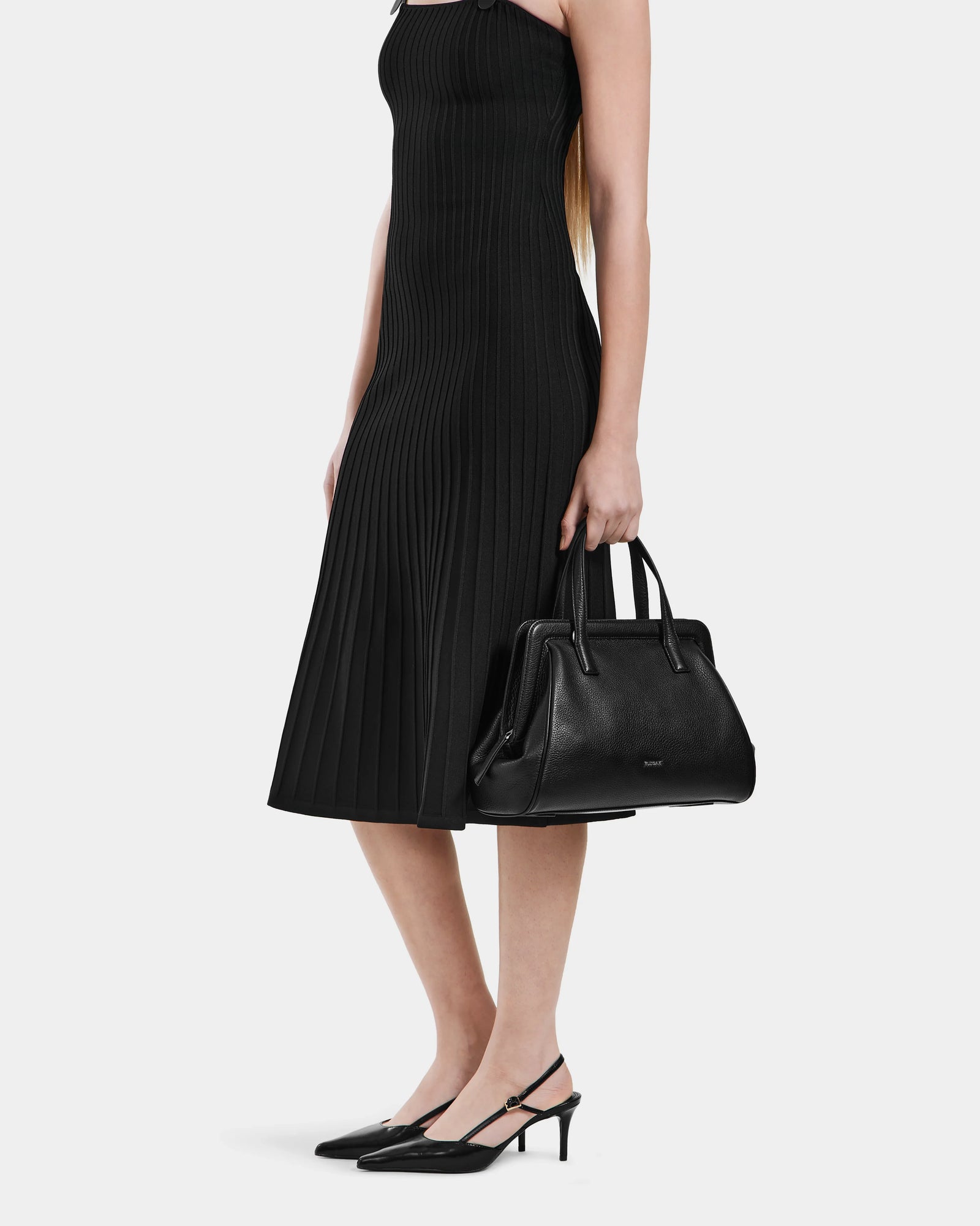 A woman in a sleeveless black dress and slingback heels holds the Rudsak TAULLA - 8325002 Black women’s pebble leather satchel, known for its spacious interior, against a plain white background.