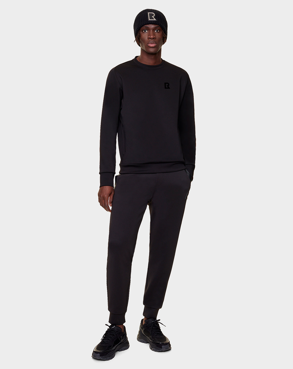 A person faces forward against a light gray background, wearing the Rudsak DEVAN - 9522510 BLACK MEN jogger pants with a black beanie, relaxed fit sweater, and black sneakers. Their left hand is in their pocket.