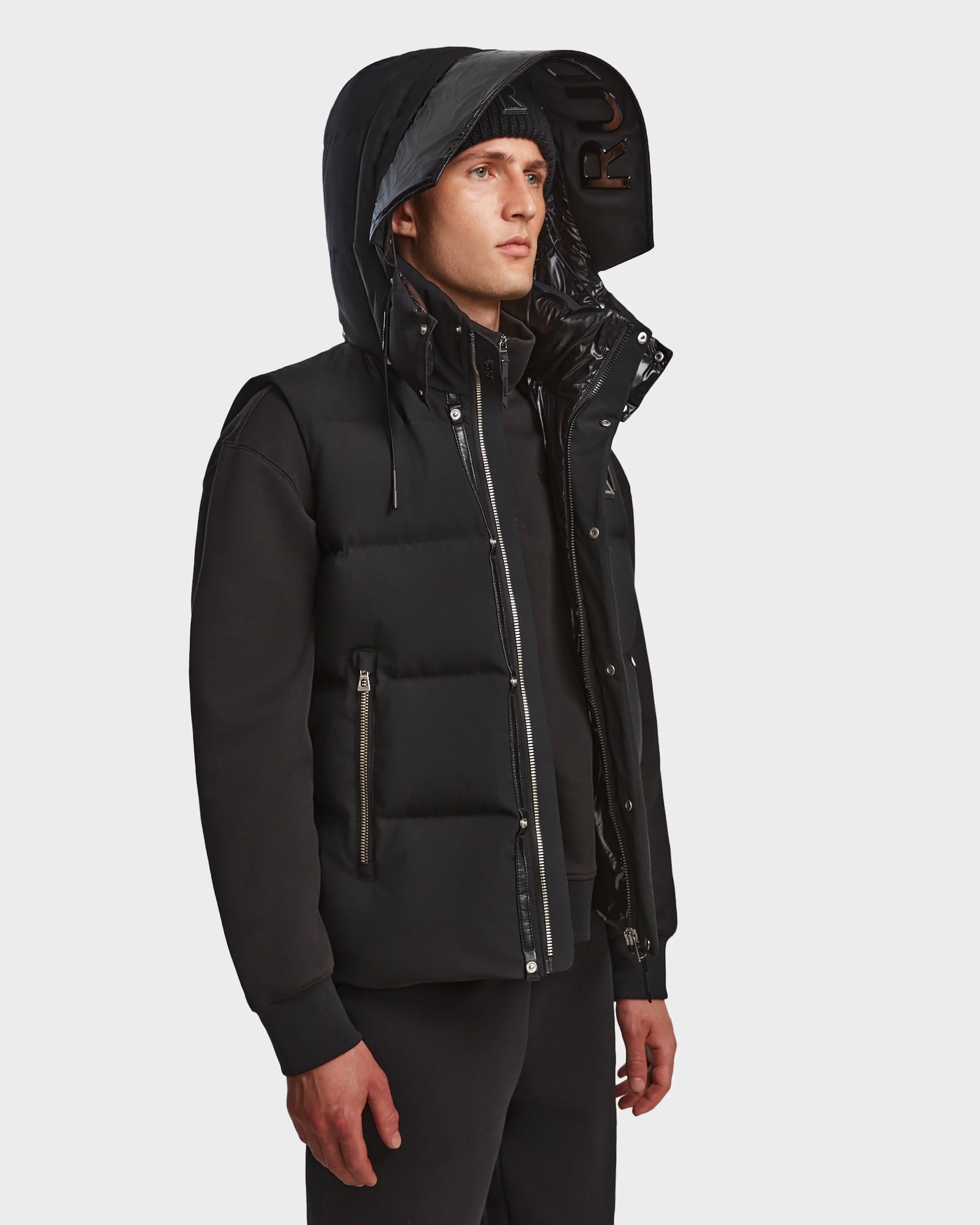 A man wears the Rudsak REN M - 9123512 Black M weather-resistant hooded puffer vest over a black sweatshirt and pants, standing slightly to the side against a plain white background.