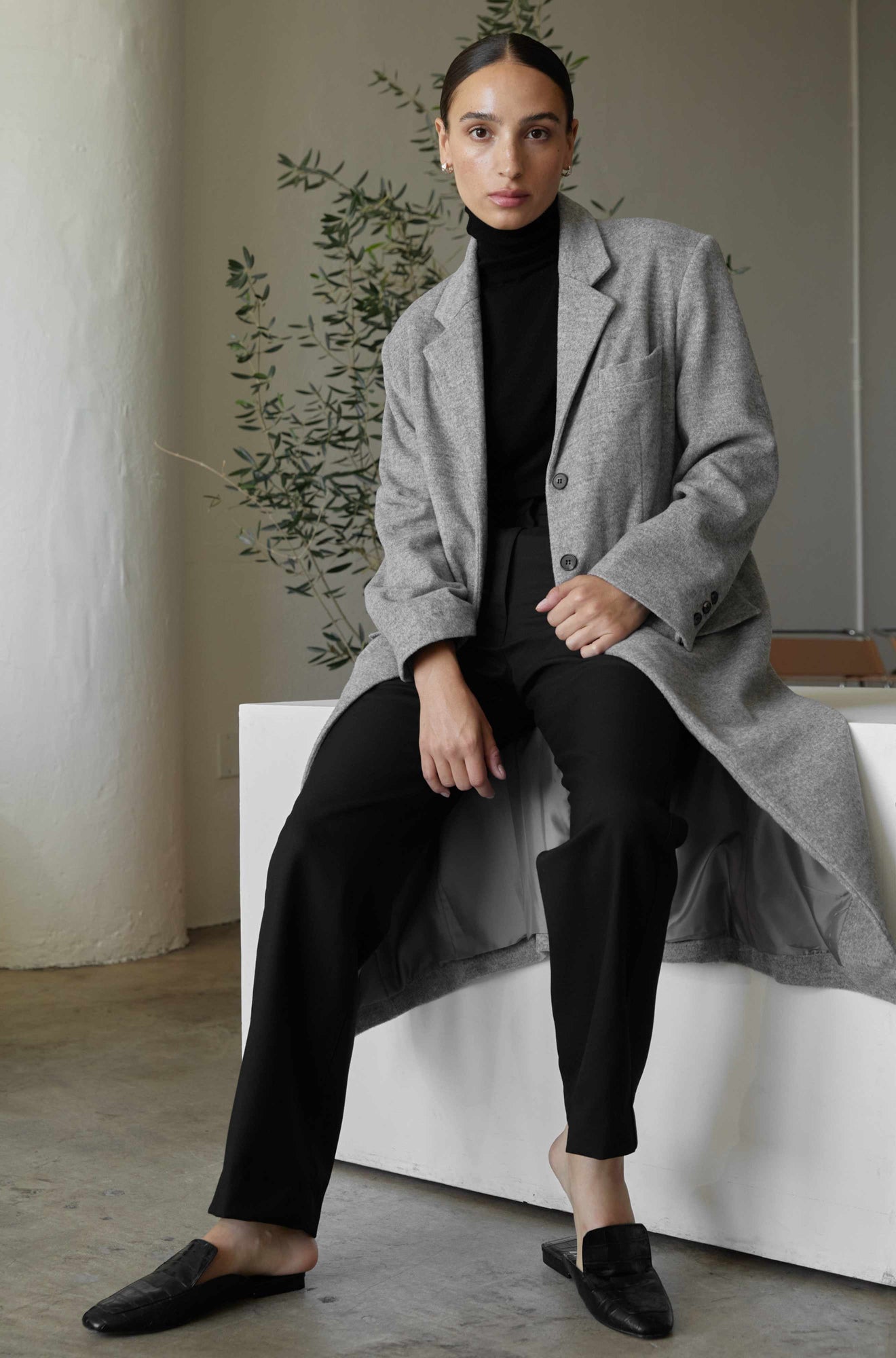 Woman wearing a gray coat over a black outfit, sitting on a white bench.