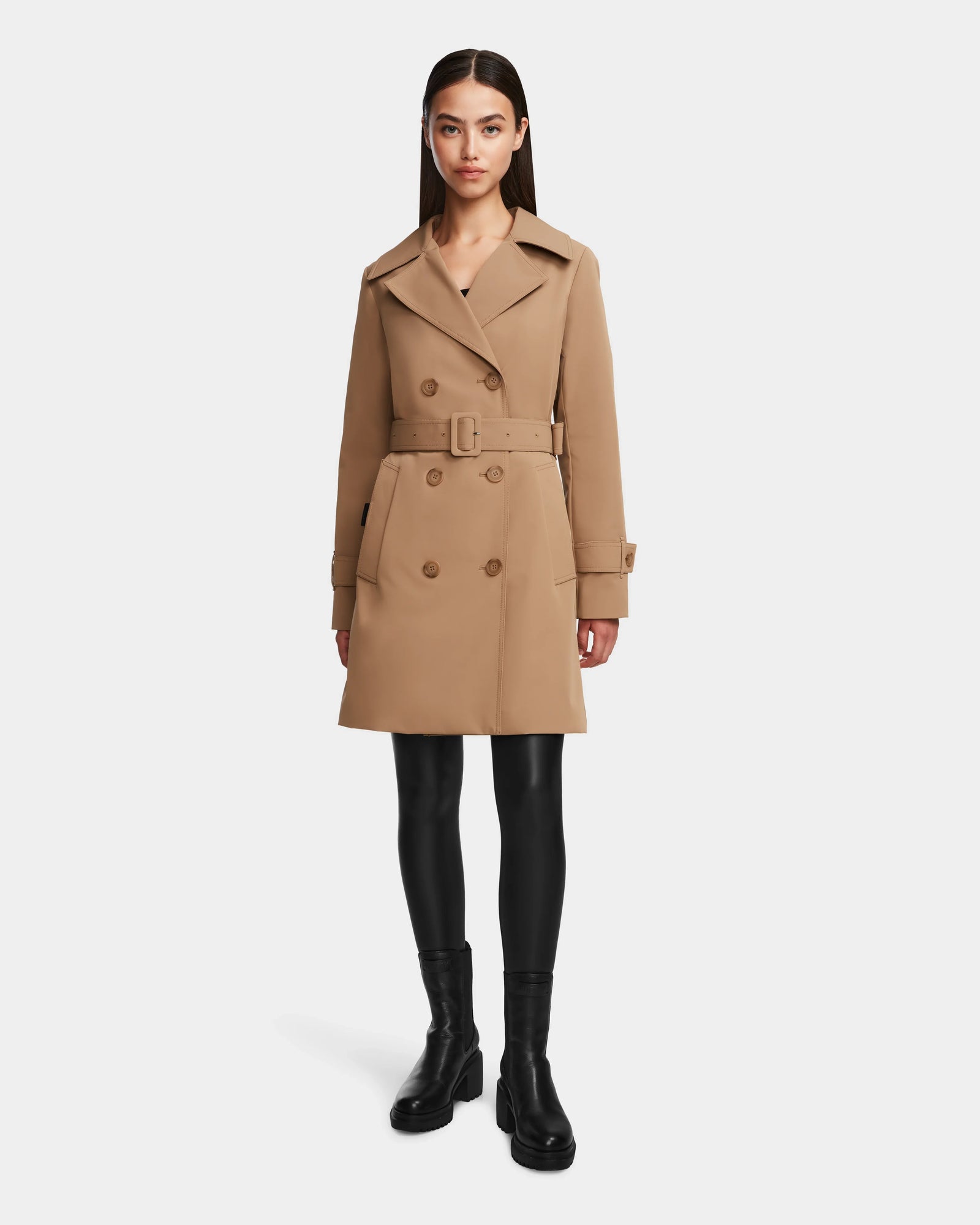 A woman faces forward against a white background, wearing the Rudsak KIMBERLY - 8124801 Camel belted double-breasted trench coat, black leggings, and black ankle boots.