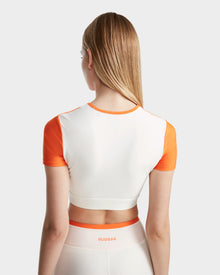 A woman with straight, light brown hair is seen from behind wearing the Rudsak LILIANE - 8524018 Eggshell fitted crop top and matching high-waisted leggings in white and orange, featuring stretch fabric and RUDSAK branding on the waistband.