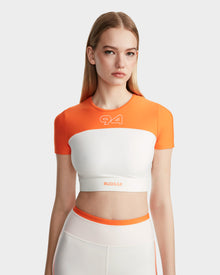 A young woman with straight, blonde hair models the Rudsak LILIANE - 8524018 Eggshell women’s fitted crop top in orange and white with 94 and RUDSAK, made from high-performance stretch fabric, paired with matching high-waisted leggings.