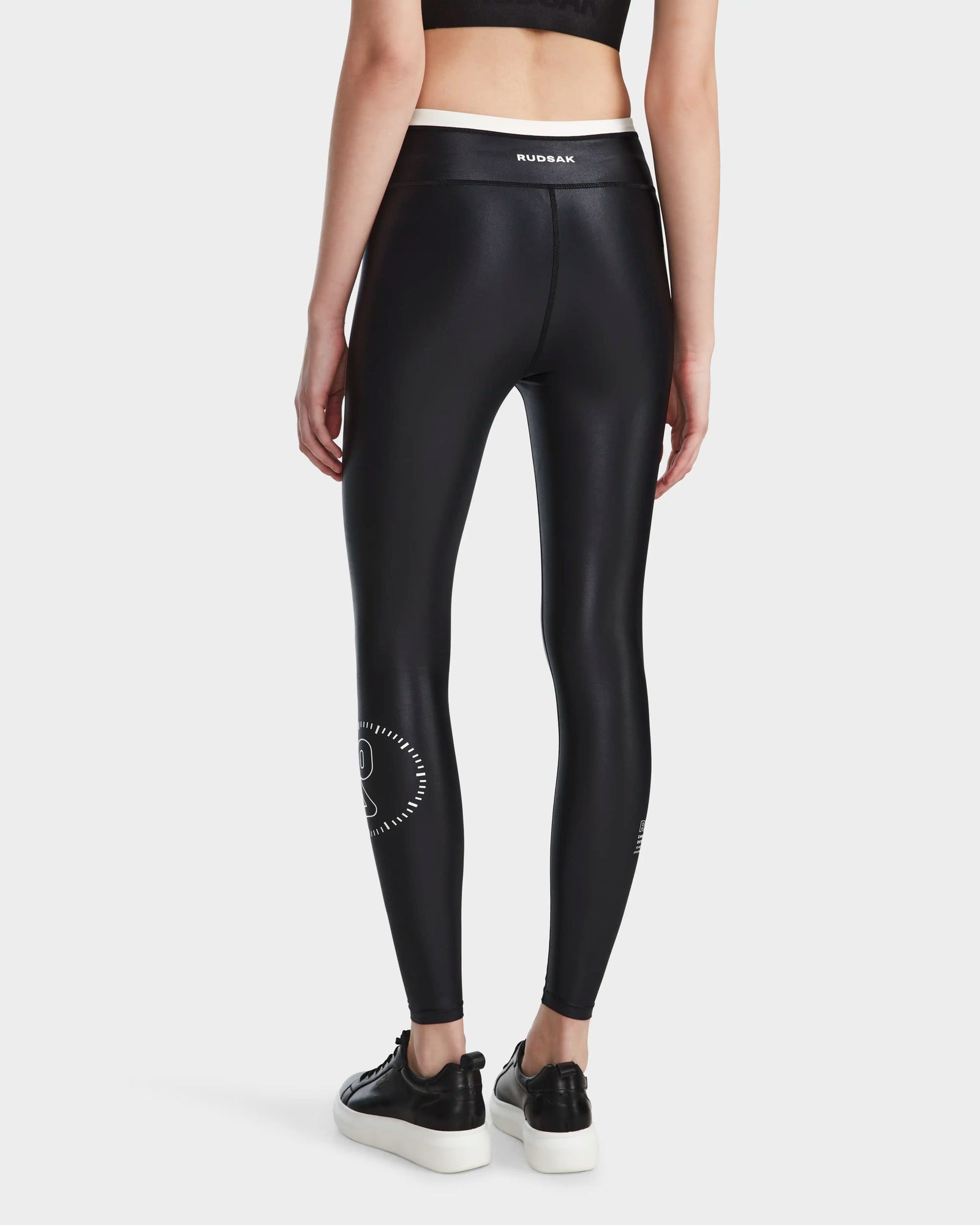 A person faces away, wearing RUDSAK TAYLA - 8524015 Black quick-dry leggings with a white design by the left calf, paired with a black crop top and black-and-white sneakers. Rudsak appears on the waistband.