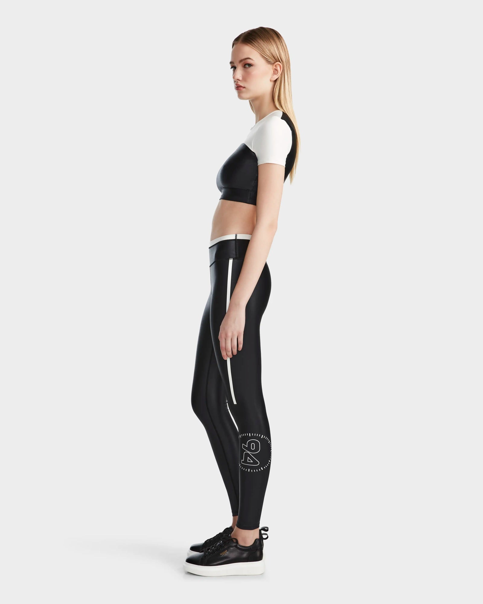 A woman with long blonde hair stands sideways, wearing the Rudsak TAYLA - 8524015 Black short-sleeve crop top, high-performance leggings with a white logo on the calf, and black platform sneakers against a plain light background.