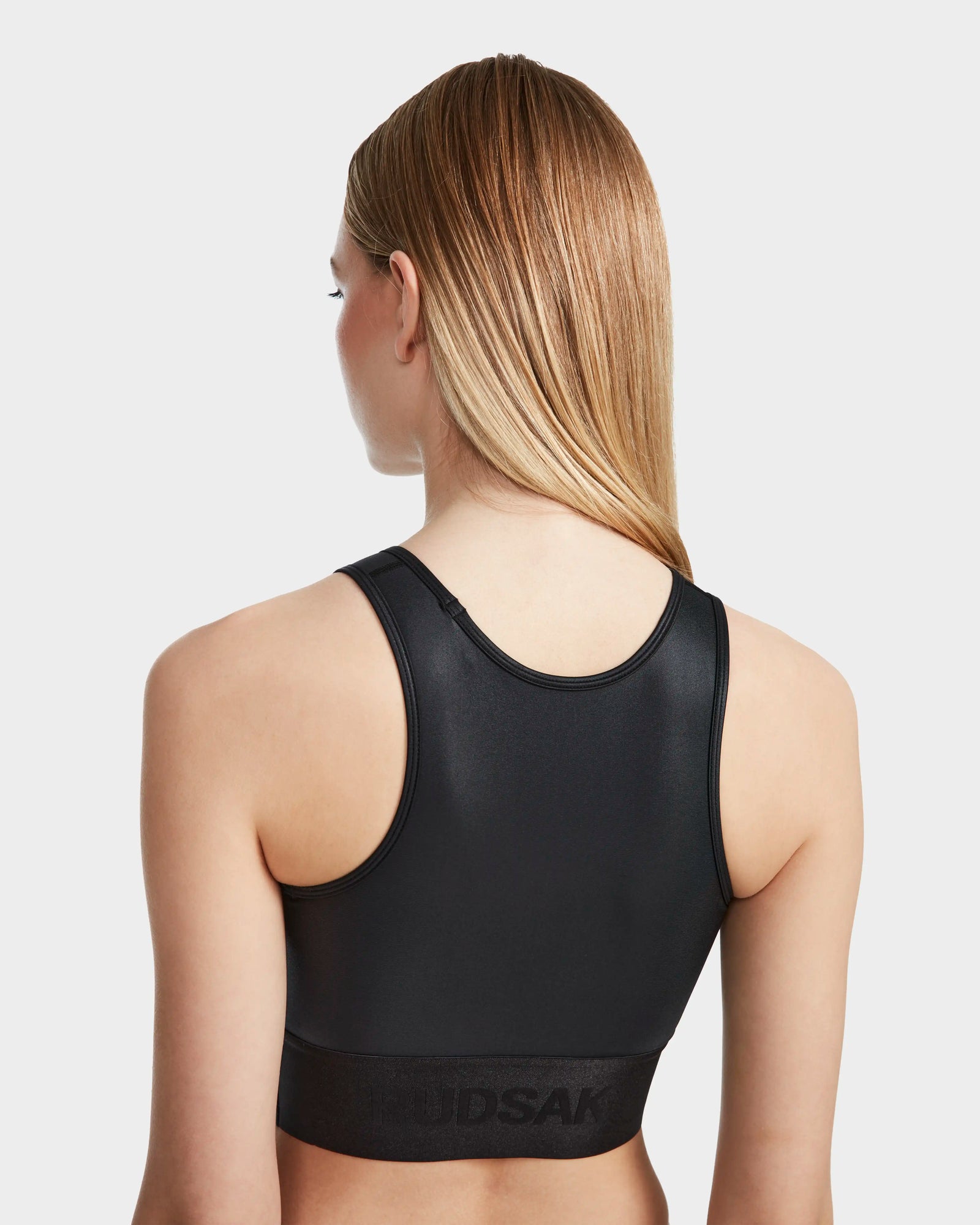 A woman with straight blonde hair is shown from the back wearing the Rudsak TRACY - 8524016 Black womens high-performance racer-back sports bra with a wide band, against a plain white background.