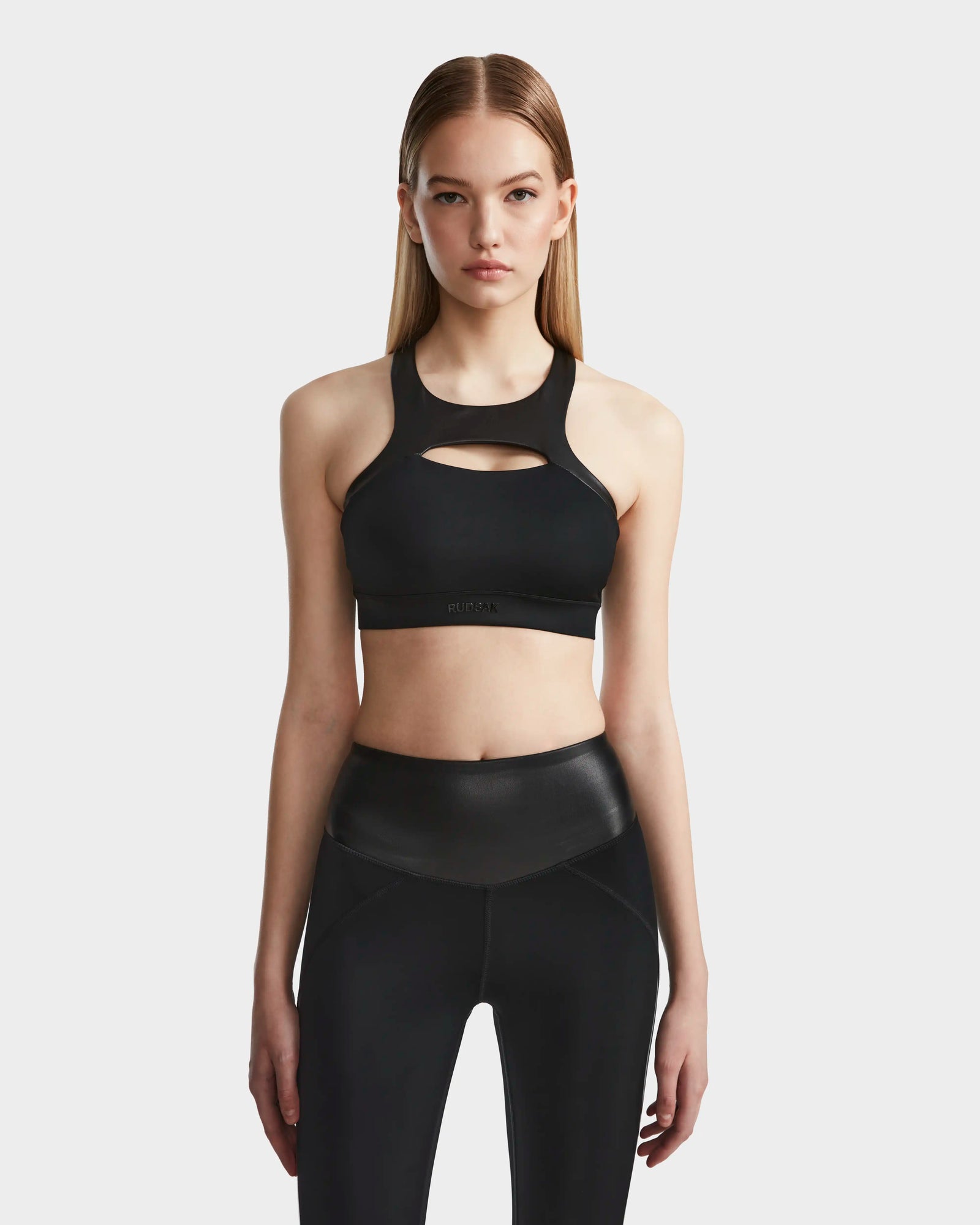 A woman with straight, long blonde hair wears the RUDSAK ARYA - 8523002 BLACK sports bra and matching high-waisted leggings, standing against a plain light gray background.