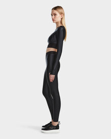 A woman with long blonde hair stands in profile, wearing the Rudsak LORA - 8524026 Black activewear top and matching high-waisted leggings with black sneakers, set against a plain light gray background.