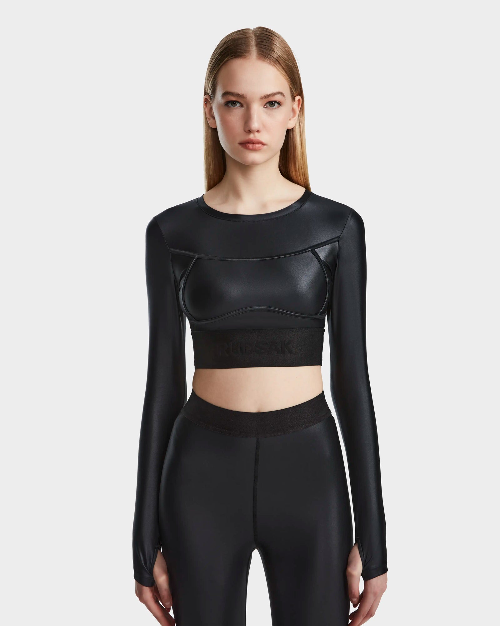 A woman with straight, light brown hair wears the Rudsak LORA - 8524026 Black fitted long sleeve top and matching high-waisted leggings, standing against a plain white background.