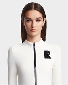 A woman with straight brown hair wears the sleek white Rudsak DAVINA - 8523534 Iceland apres-ski outfit, featuring a black zipper and LOVE brooch, standing against a plain light background and looking directly at the camera.