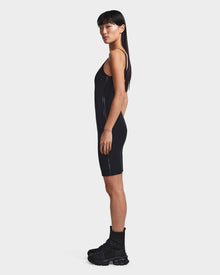 A woman in profile wears the Rudsak MILEY - 8523034 BLACK sleeveless dress with black combat boots and socks. She has long straight hair, a neutral expression, and is posed against a plain white background.