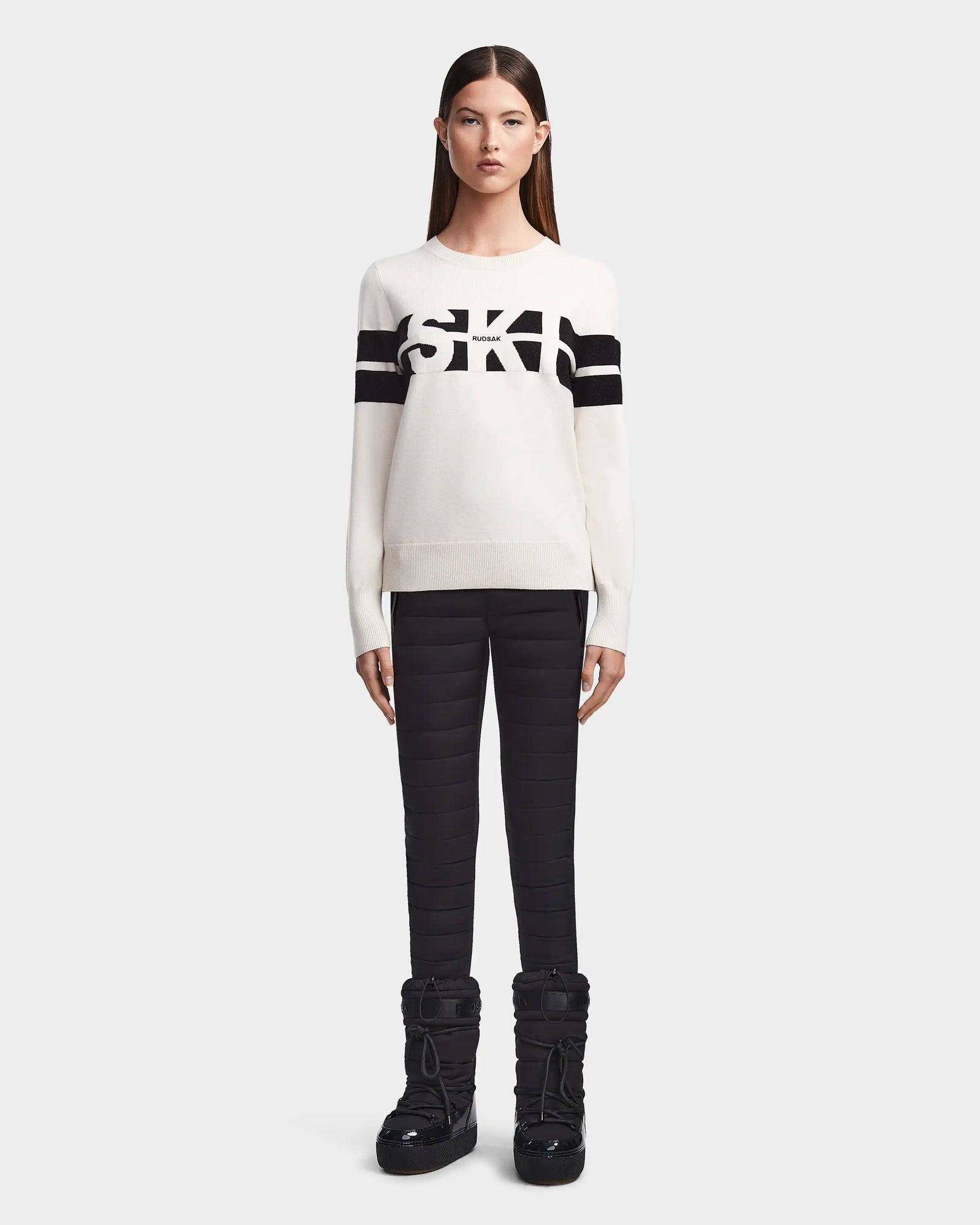 A woman faces forward in Rudsaks ODESSA - 8523540 Iceland soft-knit après-ski sweater with black stripes and “SKI” across the front, paired with black quilted pants and snow boots—ideal winter warmth.