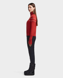A woman stands in profile wearing the Rudsak ERIKA - 8523536 Orange apres-ski turtleneck sweater with white stripes, styled with black fitted pants and wedge boots, set against a plain light gray background.