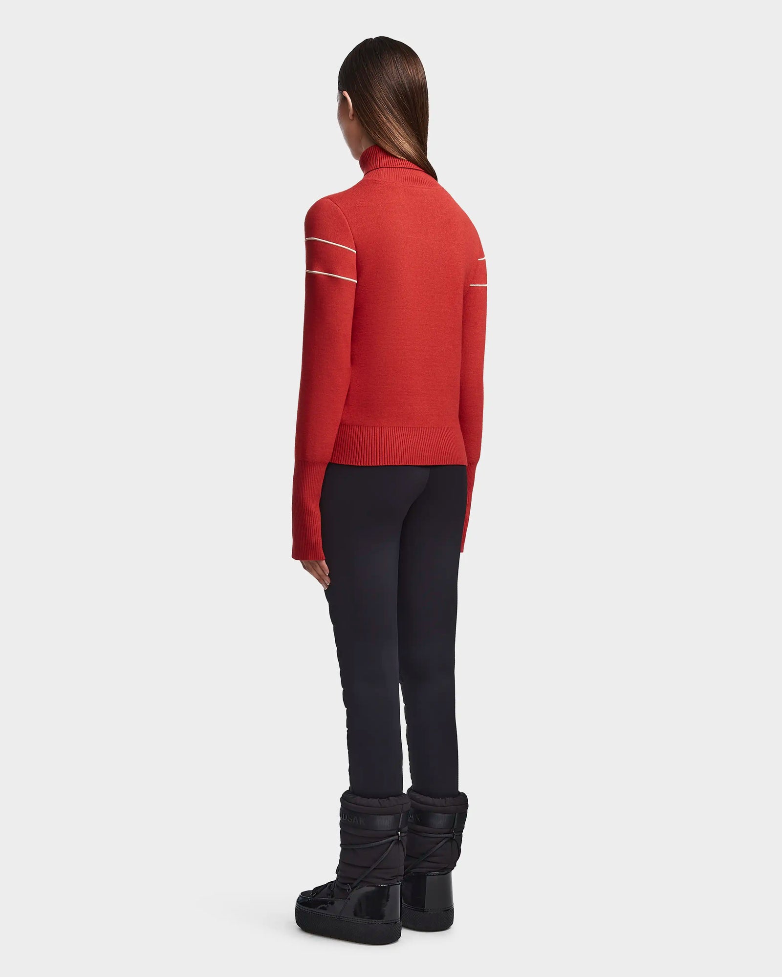 A woman stands facing away, wearing the Rudsak ERIKA - 8523536 Orange après-ski turtleneck sweater with white shoulder stripes, black fitted pants, and black winter boots. Her straight brown hair is down against a plain white background.