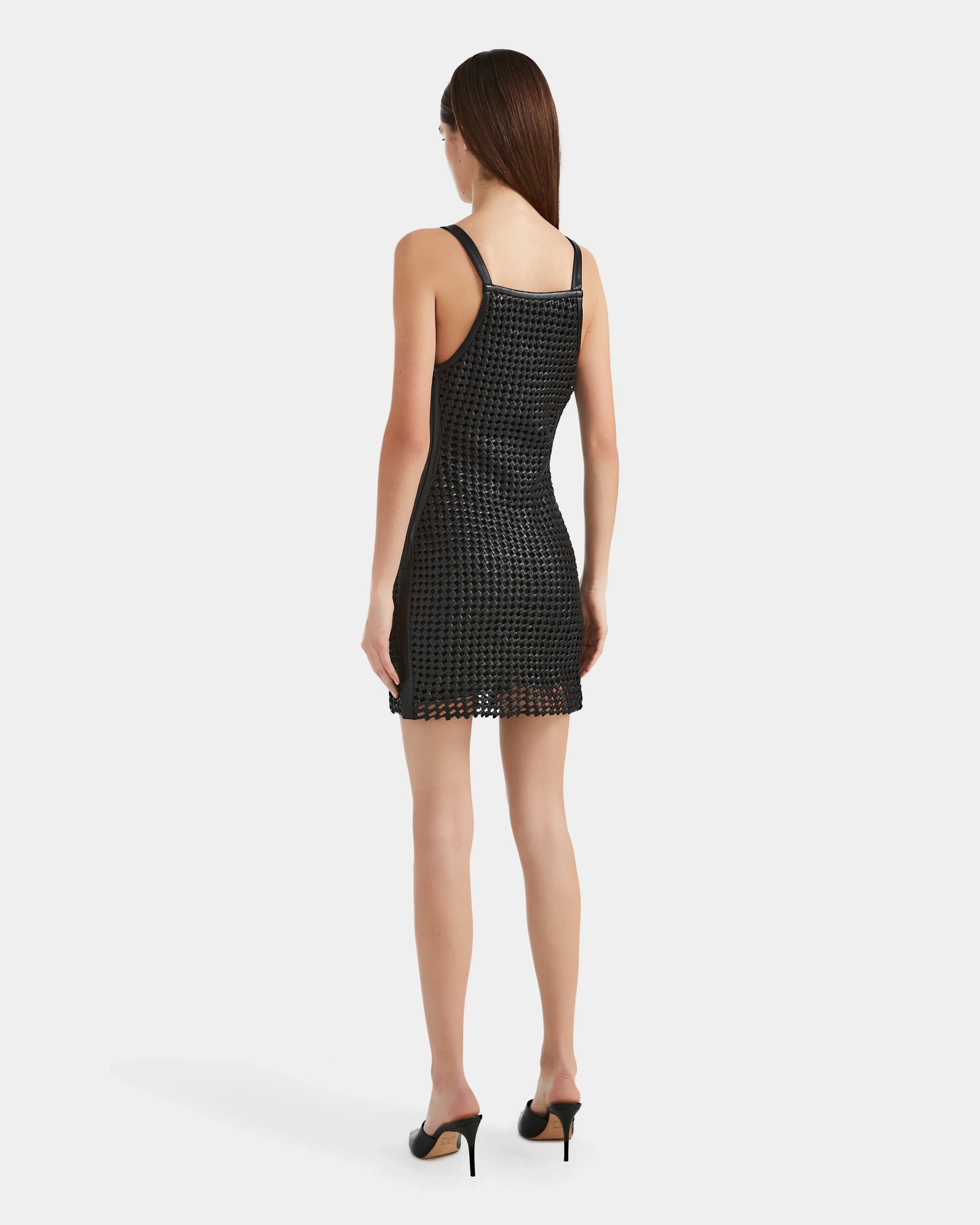 A woman with straight brown hair models the Rudsak NATALIA - 8524045 Black dress, a sleeveless fitted mesh style, paired with black high-heeled sandals, standing with her back to the camera against a plain white background.