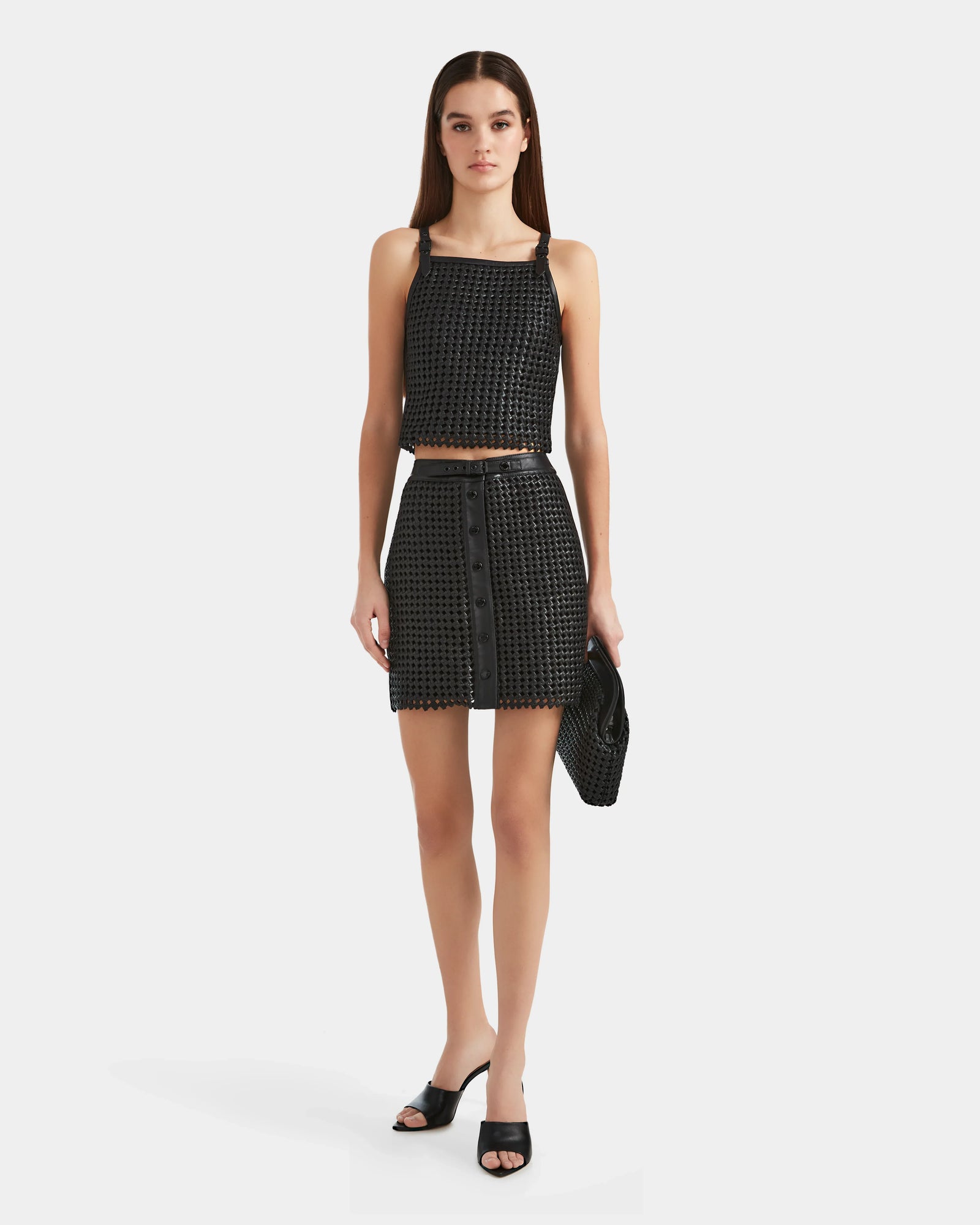 A woman stands against a white background wearing a handwoven cropped top, matching high-waisted skirt, and holding the Rudsak NADINE - 8524042 Black textured clutch—paired with black open-toe heels for a chic, luxury look.