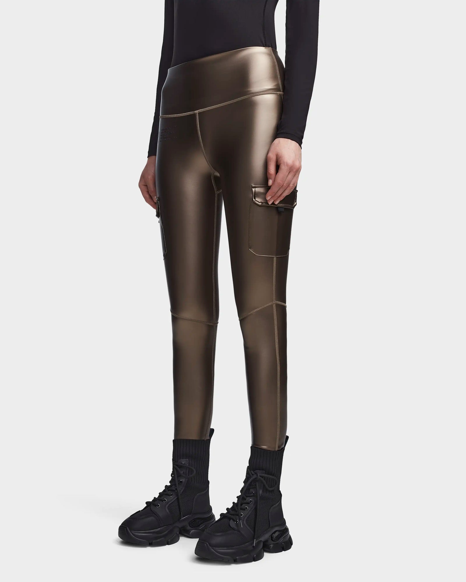 A person wearing Rudsaks SIEN P - 8523027 METALLIC CHOCOLATE stretch vegan leather cargo leggings with side pockets, a black long-sleeve top, and black high-top sneakers, standing against a plain white background.