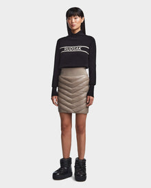 A woman faces forward wearing a black Rudsak turtleneck, the MARZI - 8523528 Cement beige chevron quilted winter skirt, and black shiny winter boots against a plain light background.