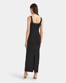 A woman stands facing away, exuding elegance in the Rudsak WANDA - 8524035 Black fitted, sleeveless maxi dress with a scoop back and hem slit, paired with white high heels.