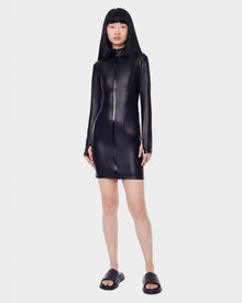 A woman with straight black hair and bangs wears the Rudsak SHANA - 8522508 BLACK long-sleeve stretch vegan leather zip-up mini dress with black slide sandals, standing against a plain white background.