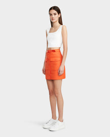 A woman faces to the side wearing Rudsaks SABINE - 8524063 Orange Sorbet quilted short skirt with large pockets, paired with a white sleeveless crop top and sneakers. A plain light gray background adds a modern touch.