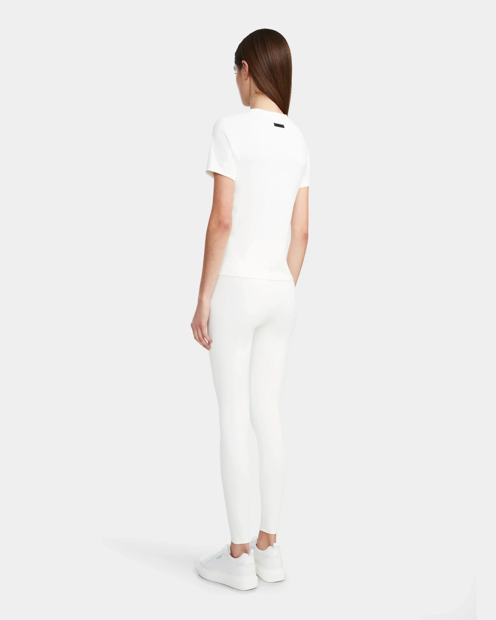 A woman with straight brown hair stands facing away on a light gray background, wearing the Rudsak SUMI - 8524030 Cotton White womens essential tee, paired with white leggings and white sneakers.