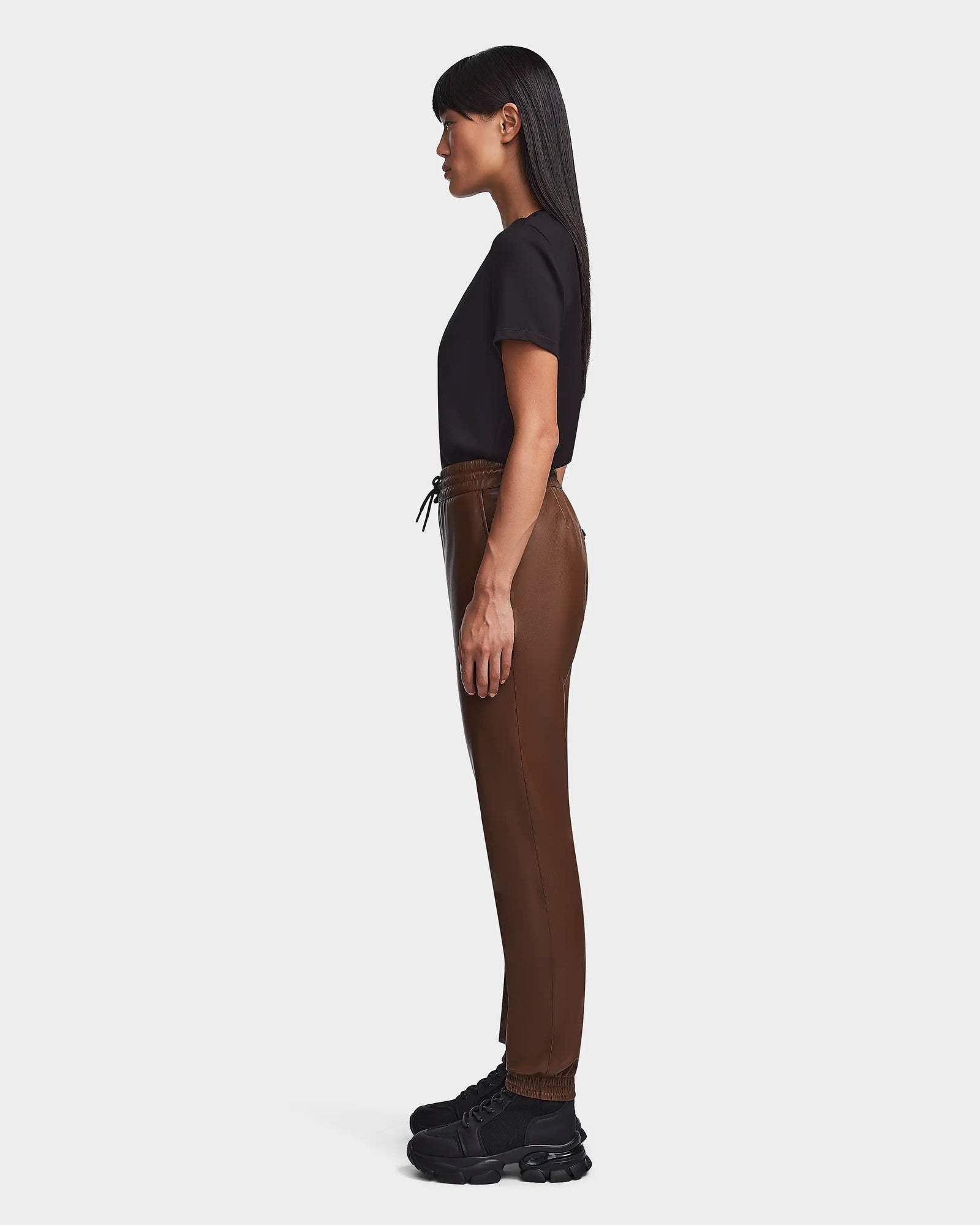 A woman stands in profile against a plain light background, wearing Rudsak's FELICIA - 8523025 COHIBA brown joggers with a drawstring, a black short-sleeve top, and black chunky sneakers.