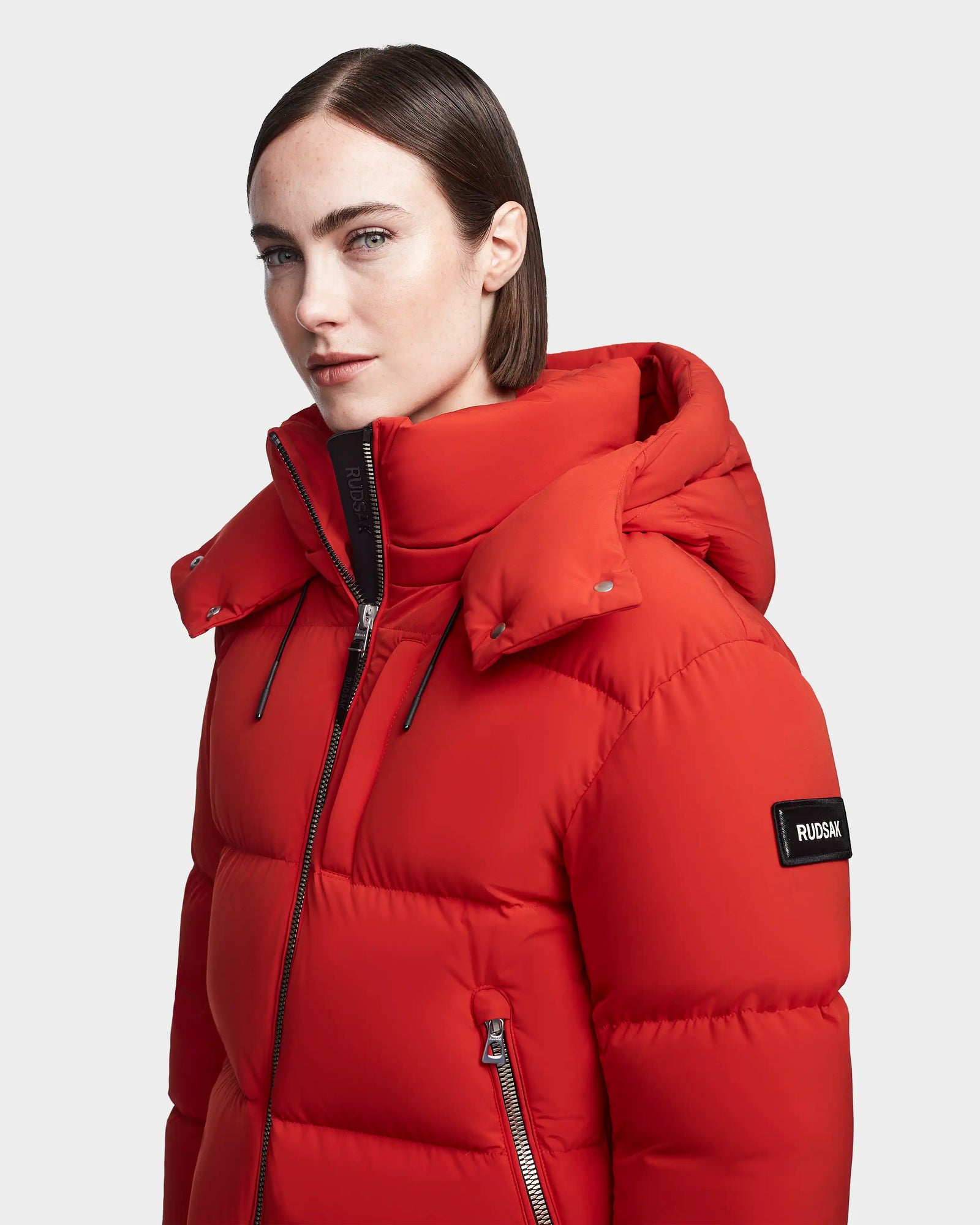 A person with straight brown hair wears a zipped-up orange Rudsak MAVE M - 8123572 womens down puffer, featuring lightweight insulation and a black “RUDSAK” patch on the sleeve. The background is plain light gray.