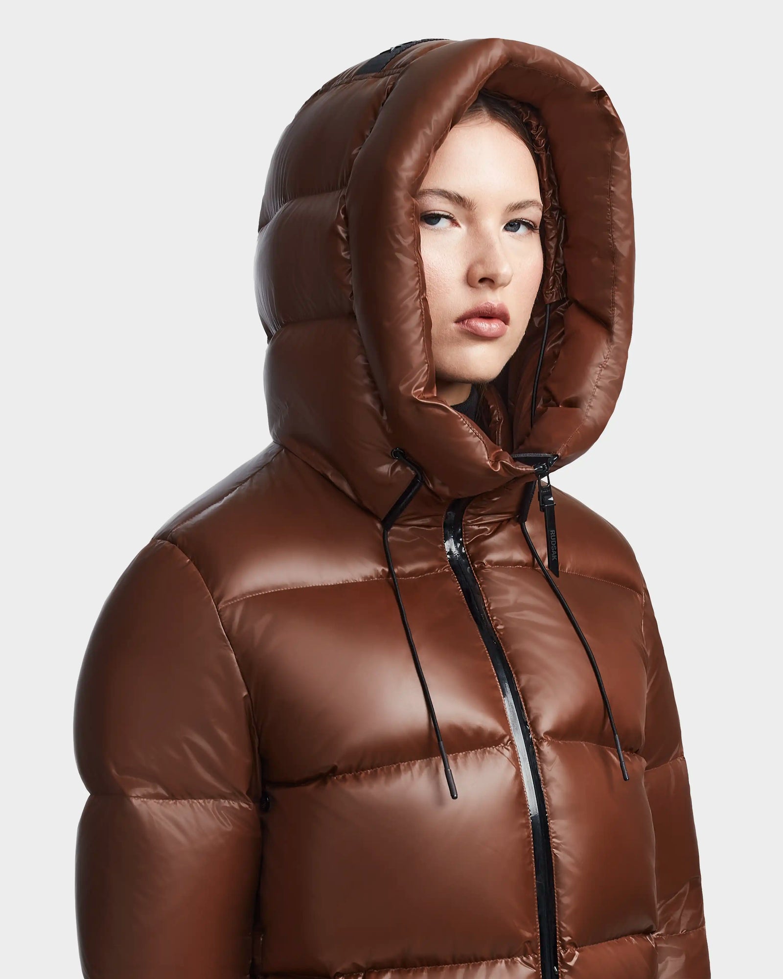 A person wearing the Rudsak RITA S - L123527 Cohiba, a shiny brown womens hooded down puffer with a large hood, looks slightly to the side against a plain light background.