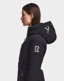 A woman with straight brown hair models the RUDSAK MILENA - 8122536 BLACK, a long black hooded down parka with bold white lettering, a white R on the upper sleeve, and recycled Stormshell fabric. Plain light gray background.