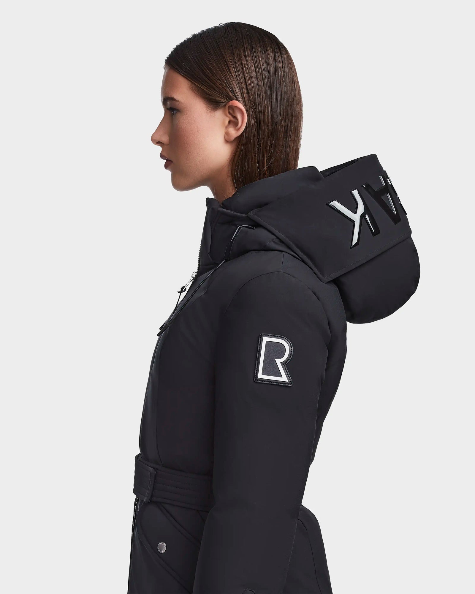 A woman with straight brown hair models the RUDSAK MILENA - 8122536 BLACK, a long black hooded down parka with bold white lettering, a white R on the upper sleeve, and recycled Stormshell fabric. Plain light gray background.
