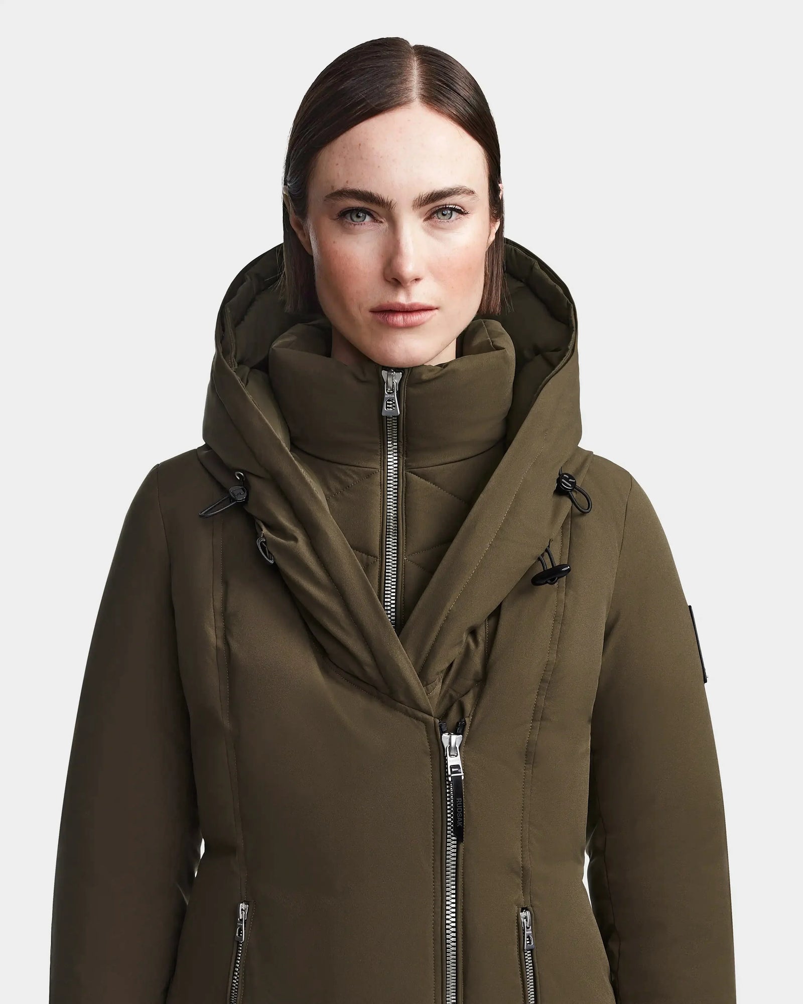 A woman with straight brown hair wears the Rudsak MAIA M - 8123582 Army jacket, featuring a high collar and multiple zippers, against a plain white background.