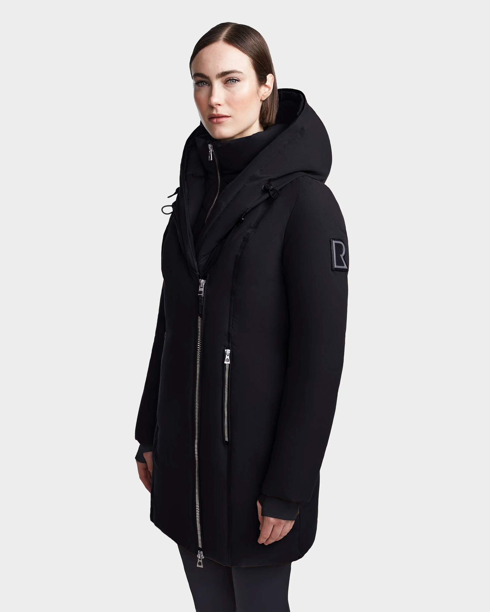A woman with straight brown hair wears the Rudsak MAIA M - 8123582 Black down parka featuring a large collar, faux fur hood, and visible zippers as she stands against a plain light gray background.