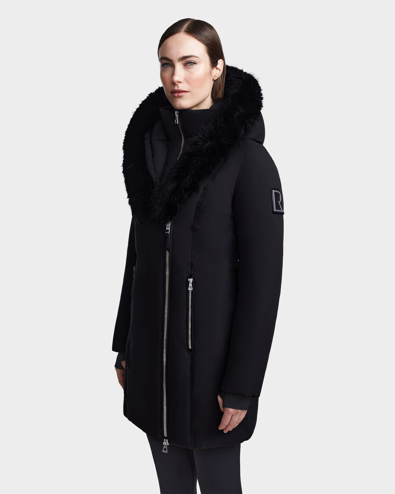 A woman wears the Rudsak MAIA M - 8123582 Black down parka with a large faux fur hood, standing facing forward with hands at her sides against a plain light gray background.