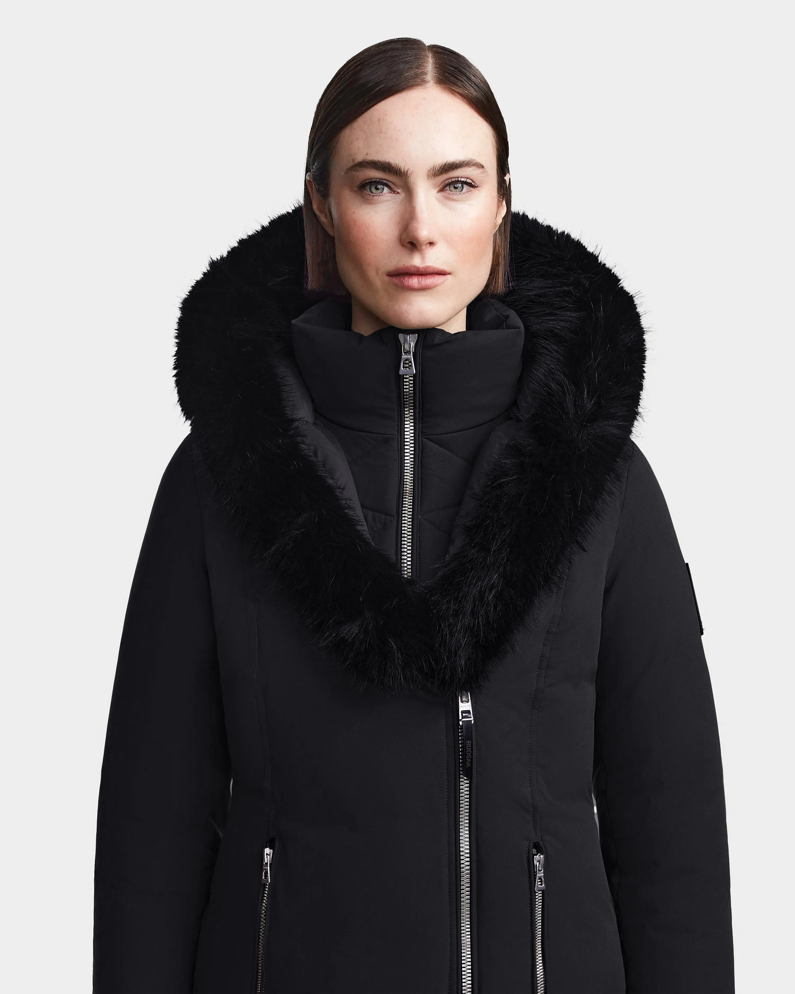 A woman wears the Rudsak MAIA M - 8123582 Black winter jacket, featuring a large, fluffy faux fur hood, standing against a plain white background.