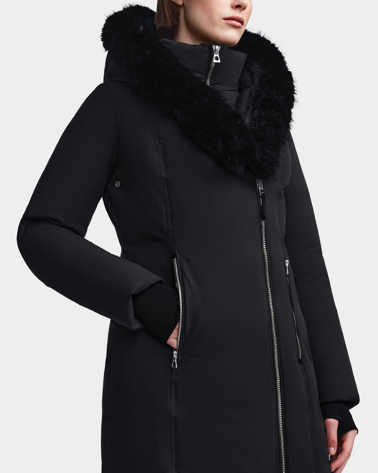 A person is wearing the Rudsak MAIA M - 8123582 Black womens down parka, featuring a large black faux fur hood and an asymmetrical front zipper. Only their lower face and upper body are visible.
