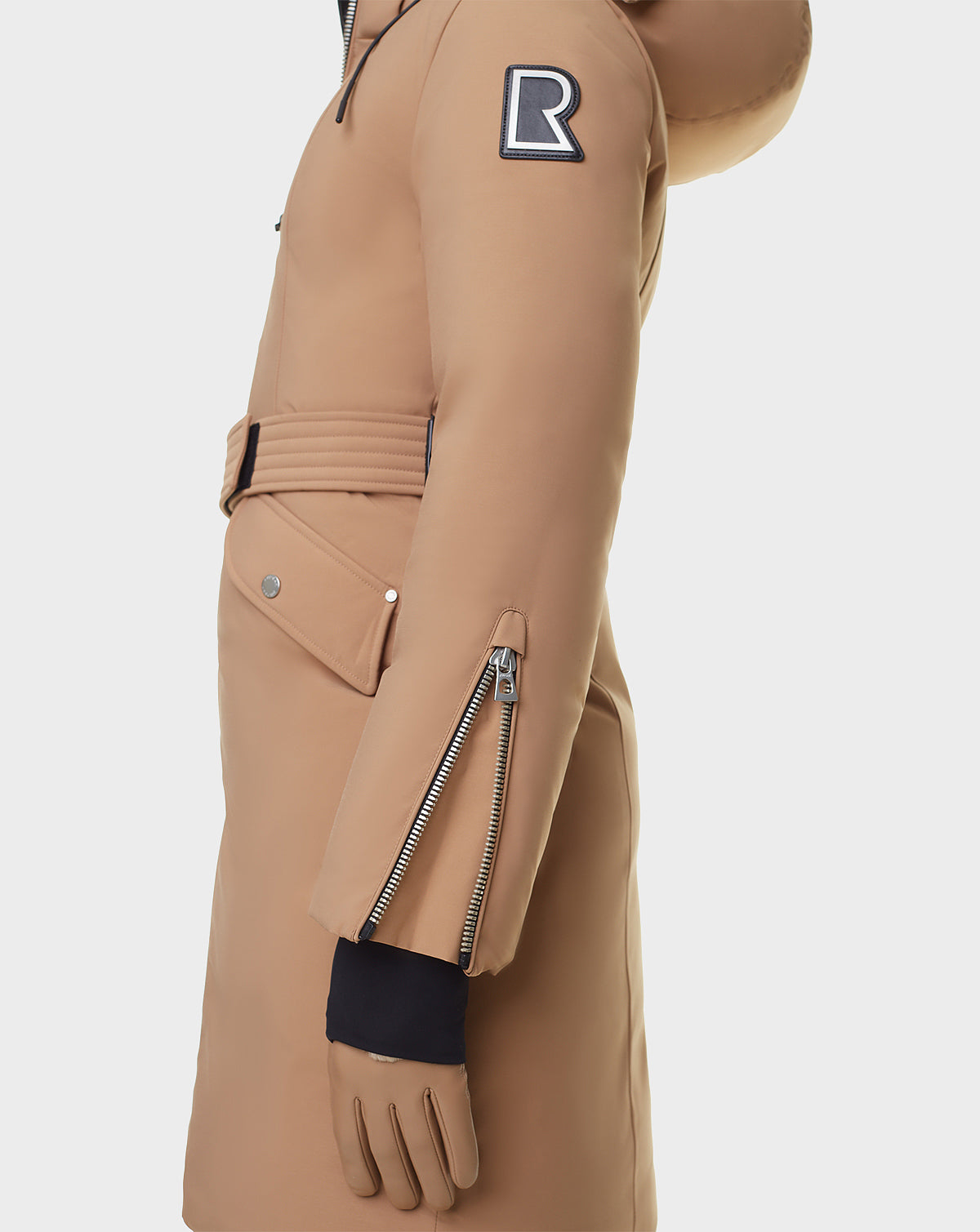 A person wearing the RUDSAK MILENA - 8122536 TOFFEE long down parka, a womens winter coat with a belted waist and large R patch on the upper arm, stands against a white background.