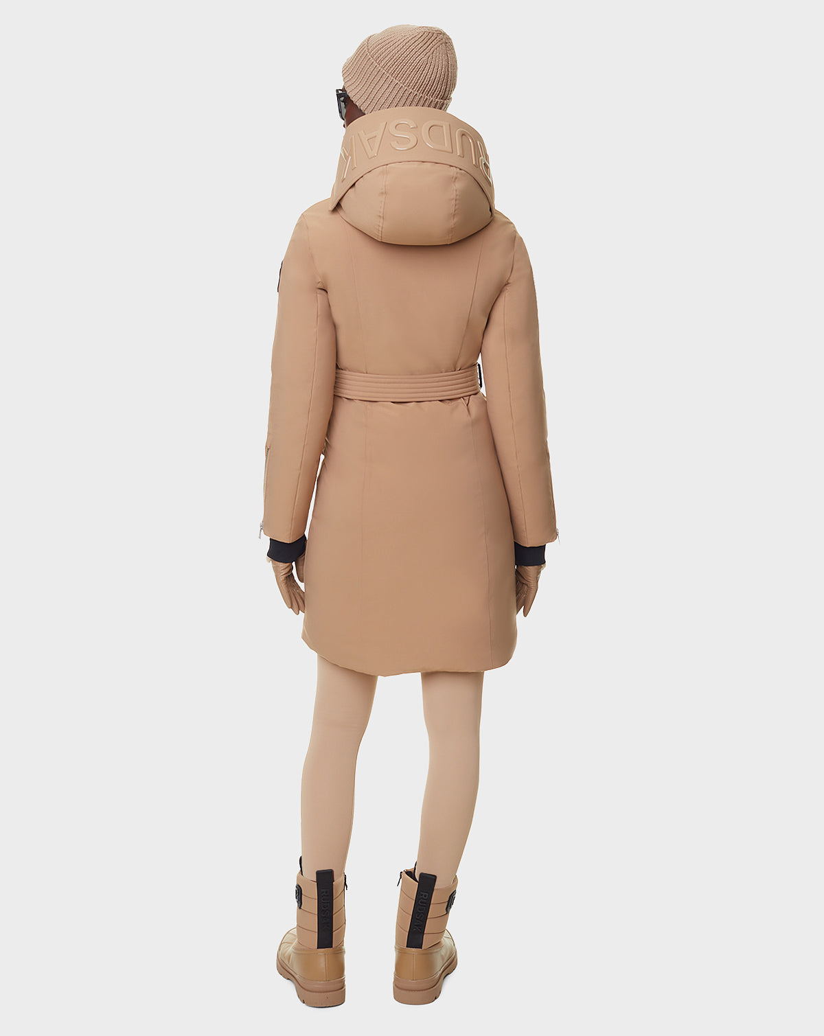 A woman stands with her back turned, wearing the RUDSAK MILENA - 8122536 TOFFEE long down parka featuring a large hood with SAVSAD text. She pairs it with tan leggings, boots, gloves, and a beanie. The background is plain white.