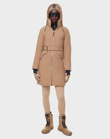 A person wearing the RUDSAK MILENA - 8122536 TOFFEE long down parka with a belt, tan leggings, tan lace-up boots, black gloves, black sunglasses, and a tan cap with a black R logo stands against a plain white background.