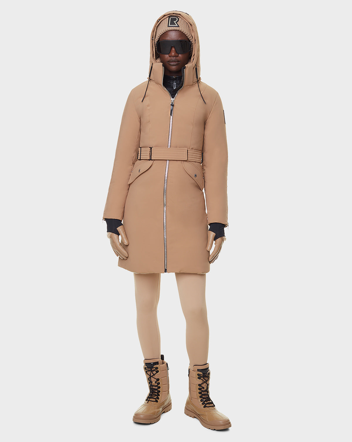 A person wearing the RUDSAK MILENA - 8122536 TOFFEE long down parka with a belt, tan leggings, tan lace-up boots, black gloves, black sunglasses, and a tan cap with a black R logo stands against a plain white background.