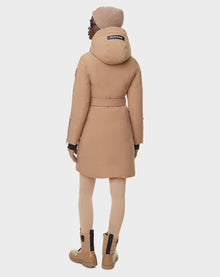 A person in the RUDSAK MILENA - 8122536 TOFFEE womens long down parka, beige with matching knit hat, leggings, and boots, stands facing away on a white background. The hood displays the visible RUDSAK logo.