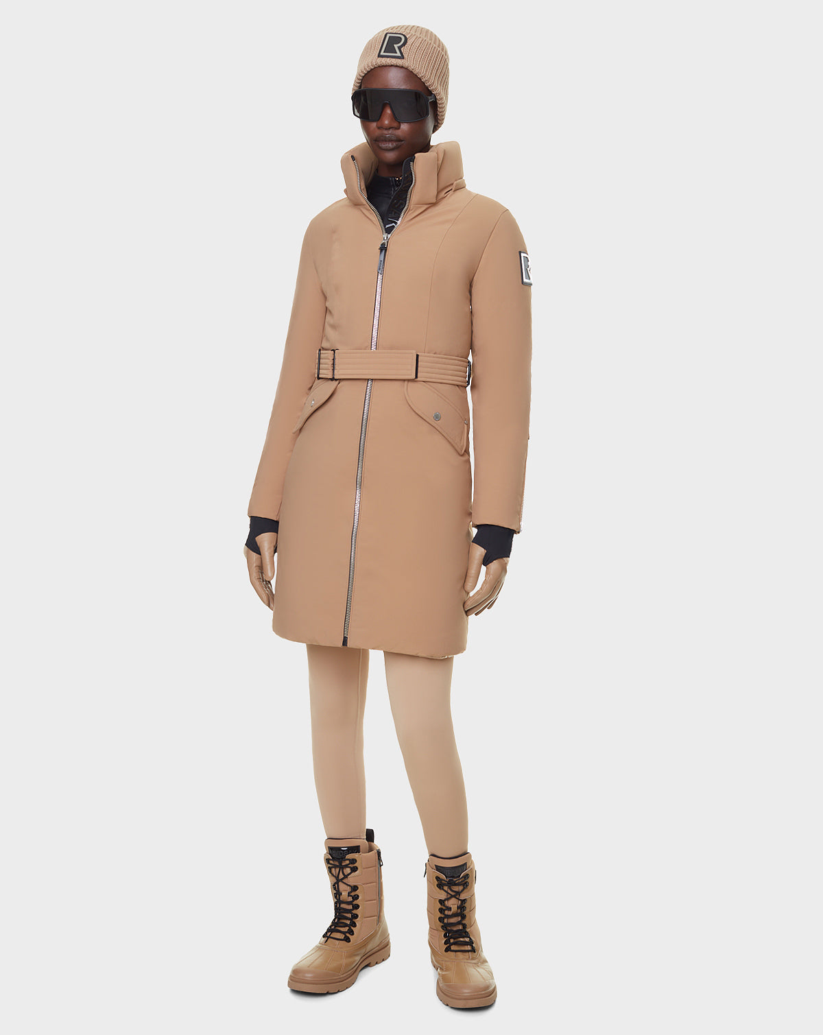 A person stands against a white background wearing RUDSAK’s MILENA - 8122536 TOFFEE women’s winter coat, a matching beanie, tan leggings and lace-up boots, black gloves, and oversized black sunglasses.