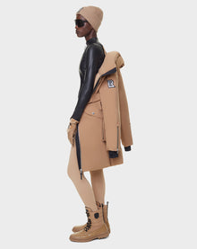 A person in a black bodysuit, tan beanie, RUDSAK MILENA - 8122536 TOFFEE coat draped over their shoulders, tan gloves, leggings, boots, and sunglasses stands in profile against a plain white background.